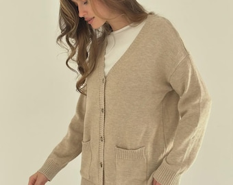 May include: A beige cardigan sweater with a V-neck, button closure, and two front pockets. The sweater has ribbed cuffs and a ribbed hem. The person is wearing a white shirt underneath and black pants.