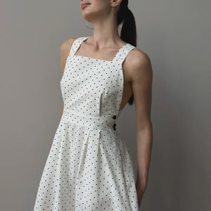 Puede incluir: Vestido blanco con lunares negros, con escote cuadrado y tirantes anchos. El vestido presenta un corpi&ntilde;o entallado, una cintura definida y una falda acampanada. Botones negros adornan la cintura. La tela es ligera.