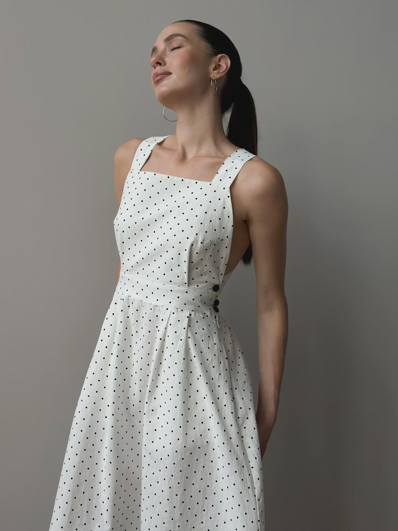 Puede incluir: Vestido blanco con lunares negros, con escote cuadrado y tirantes anchos. El vestido presenta un corpi&ntilde;o entallado, una cintura definida y una falda acampanada. Botones negros adornan la cintura. La tela es ligera.