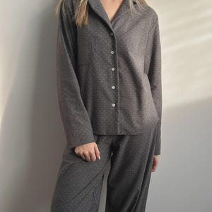 Op de afbeelding: Grijze pyjamaset met een subtiel wit stippenpatroon. De top met lange mouwen heeft een kraag, een borstzak en een knoopsluiting. De bijpassende broek heeft een losse pasvorm. De stof lijkt zacht en comfortabel.