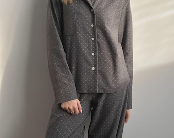 May include: Gray pajama set with a subtle white polka dot pattern. The long-sleeved top has a collared neckline, a chest pocket, and a button-down front. The matching pants are loose-fitting. The fabric appears soft and comfortable.