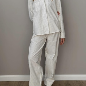 Può includere: Pigiama bianco con un piccolo motivo a pois scuri. Il set include una camicia abbottonata con taschino sul petto e pantaloni abbinati. La persona indossa pantofole grigie e soffici.