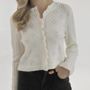 Puede incluir: Rebeca color crema con cuello festoneado y cierre de botones. El suéter de manga larga presenta un patrón calado en forma de diamante y puños acanalados. La modelo lleva vaqueros negros.