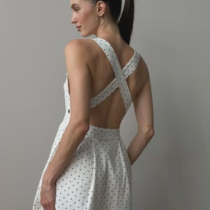 Puede incluir: Vestido blanco con lunares negros. El vestido presenta un dise&ntilde;o de espalda cruzada y una falda plisada. La modelo lleva el vestido, con una coleta y aros. Un estilo cl&aacute;sico y femenino.