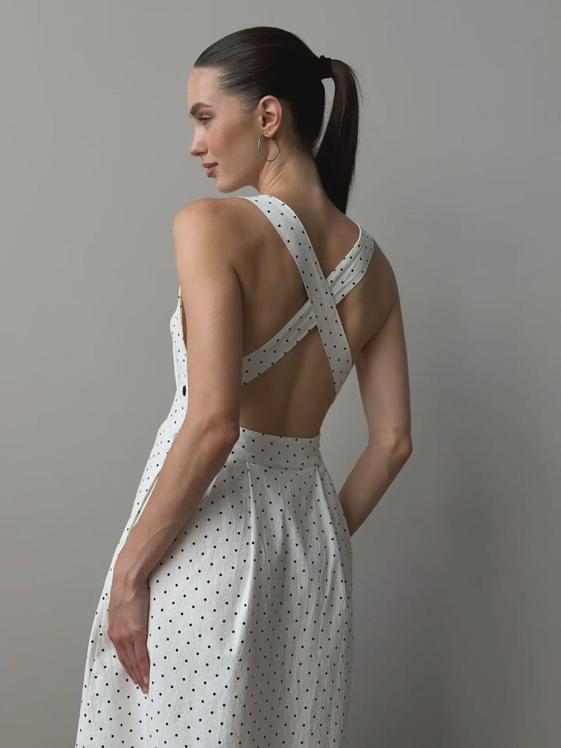 Puede incluir: Vestido blanco con lunares negros. El vestido presenta un dise&ntilde;o de espalda cruzada y una falda plisada. La modelo lleva el vestido, con una coleta y aros. Un estilo cl&aacute;sico y femenino.
