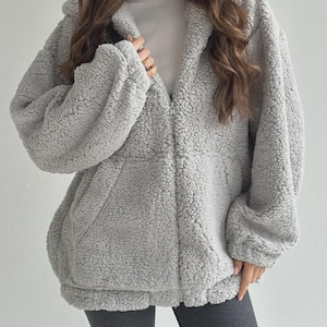 May include: A light gray, long-sleeved, zip-up jacket made of a textured, fuzzy fleece material. The jacket features a hood, a full-length front zipper, and two side pockets. It is worn over a plain white top and dark gray leggings.
