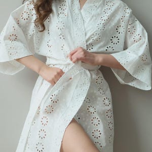 May include: White eyelet robe with a floral pattern. The robe has wide sleeves and a tie belt. The fabric is lightweight and appears to be cotton. The robe is knee-length and has a relaxed fit.