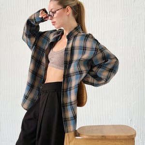 May include: A blue, tan, and black plaid shirt is worn over a gray sports bra and black pants. The person is wearing glasses and is leaning against a wooden stool. The background is a white textured wall.