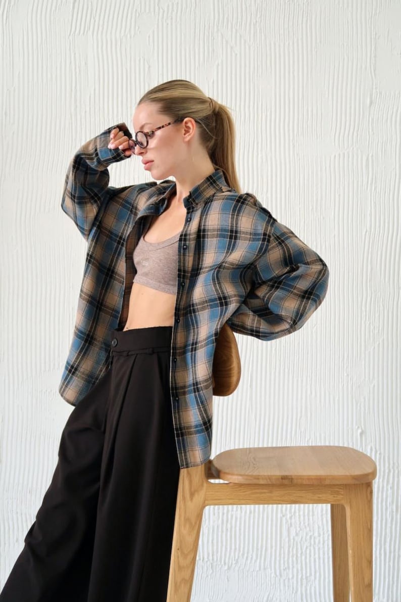 May include: A blue, tan, and black plaid shirt is worn over a gray sports bra and black pants. The person is wearing glasses and is leaning against a wooden stool. The background is a white textured wall.