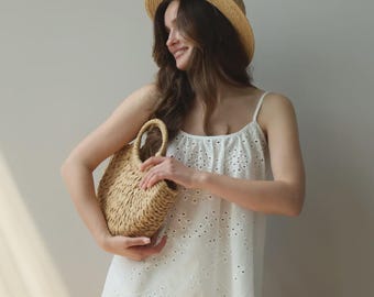 May include: A woman wearing a white eyelet top and shorts set, paired with a straw hat and a woven handbag. The outfit is light and airy, perfect for warm weather. The eyelet details add a touch of elegance to the casual ensemble.