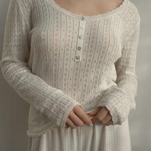 May include: Cream-coloured long-sleeved top and matching bottoms. The top features a scoop neckline, a button-up front, and a delicate floral pattern. The sleeves and hem have a scalloped edge. The bottoms are a matching cream colour.