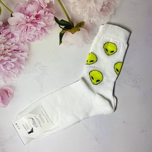 May include: White socks with a yellow alien face pattern. The socks are folded and lying on a white surface with pink flowers in the background.