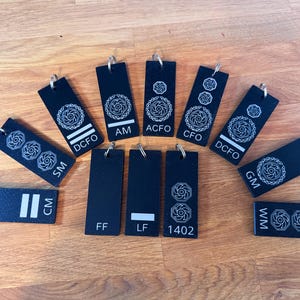May include: A selection of black rectangular keyrings with silver hoops. Each keyring displays a white, stylized floral design and various white text abbreviations, including "SM", "CM", "DCFO", "AM", "ACFO", "CFO", "GM", "WM", "FF", "LF", and "1402".
