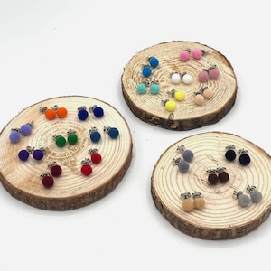 Könnte beinhalten: Eine Sammlung farbenfroher Pompon-Ohrstecker, die auf rustikalen Holzscheiben präsentiert werden. Die Ohrringe sind in verschiedenen Farben erhältlich, darunter Rot, Orange, Grün, Blau, Lila und Rosa. Die Ohrringe sind klein und rund, mit einer flauschigen Textur.
