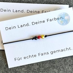 Könnte beinhalten: Ein schwarzes Kordelarmband mit drei facettierten Perlen in Schwarz, Rot und Gelb, die die deutsche Flagge darstellen. Das Armband wird auf einer Karte mit deutschem Text und einer Weltkugelgrafik präsentiert.