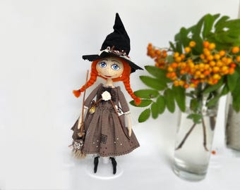 May include: A handmade witch doll with red hair, a black witch hat, and a brown plaid dress. She is holding a broom and is standing on a white base.
