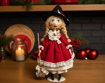May include: A handmade doll dressed in a red dress with white lace trim, a black hat, and blonde braids. The doll holds a small broom and is standing on a wooden surface. The doll is a festive holiday decoration.