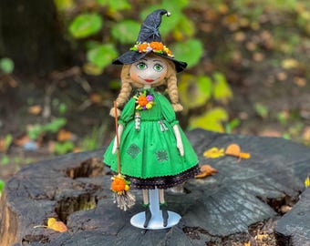 May include: A handmade witch doll with blonde hair, a green dress, and a black witch hat with orange and purple flowers. The doll is holding a broom and is standing on a white pedestal.