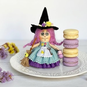 Peut inclure: Une poupée artisanale déguisée en sorcière, portant un chapeau noir orné de fleurs, une robe bleue et tenant un balai. Une pile de quatre macarons jaunes et violets est posée sur une assiette blanche. La poupée a des cheveux roses.