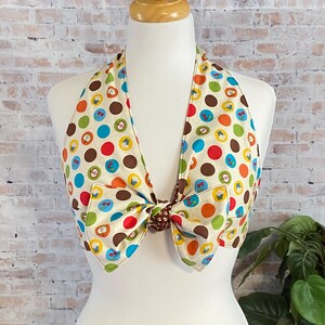May include: A halter top featuring a cream background with a colorful pattern of polka dots and small icons including apples, cherries, and cupcakes. The garment has a large fabric bow tied at the center front and a halter neck strap design.