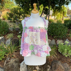 Peut inclure: Un haut sans manches en patchwork composé de divers carrés de tissu aux tons pastel, notamment lavande, rose et vert. Le panneau central présente une illustration brodée d'une personne en robe. Le haut possède des bretelles à nouer.