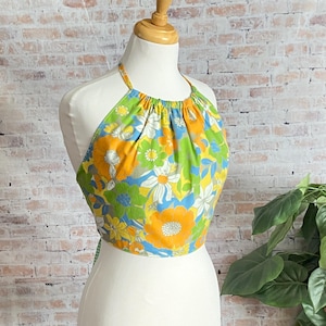 May include: A halter top with a vibrant floral pattern in yellow, orange, green, blue, and white. The top is displayed on a mannequin, showcasing its design and shape. The fabric has a gathered neckline and ties at the back.