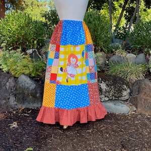Puede incluir: Una falda larga hasta los tobillos con un diseño de patchwork que presenta varios patrones coloridos, incluyendo lunares, estampados florales pequeños y un panel central amarillo con una muñeca de dibujos con pelo de lana rojo.