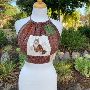 Può includere: Un top corto all'americana con un design patchwork caratterizzato da motivi floreali marroni, verdi e crema. Il pannello centrale mostra un'illustrazione dettagliata di un gatto a pelo lungo marrone e bianco seduto tra fiori su sfondo crema.