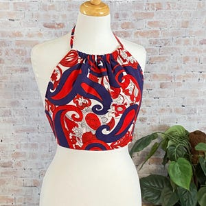 May include: A halter top with a vibrant paisley pattern in red, white, and navy blue. The top is displayed on a mannequin, showcasing its design and style. The fabric features a repeating floral and swirl design.
