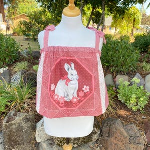 Puede incluir: Una blusa sin mangas en tonos rosa empolvado y blanco, con un panel octogonal central que muestra la ilustración de un conejo blanco con un lazo rojo, rodeado de motivos florales. La prenda tiene tirantes para anudar y cuello fruncido.