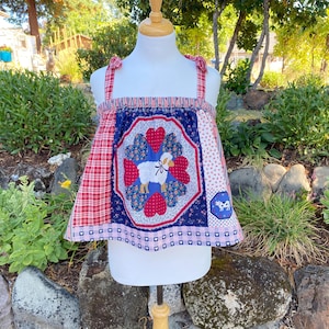 May include: A sleeveless top featuring a patchwork design with red and white plaid, floral patterns, and a central navy blue panel. The centre displays a white sheep graphic surrounded by red and blue hearts. It has tie-shoulder straps.