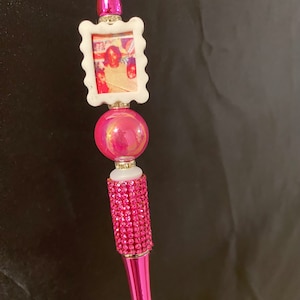 May include: A pink and white pen with a photo frame on top. The frame has a picture of a group of people. The pen is covered in rhinestones.