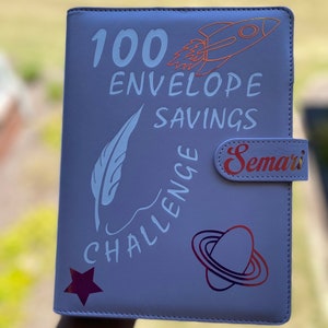 May include: A purple 100 Envelope Savings Challenge binder with a rocket, feather, star, and planet design. The text "Semari" is on the side of the binder.
