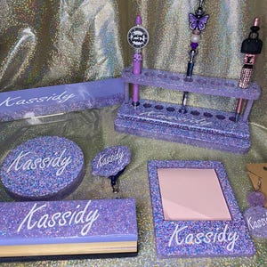 May include: A purple glitter desk organizer set with the name "Kassidy" on each piece. The set includes a pen holder, a notepad, a small round tray, a small square tray, and a pair of earrings.
