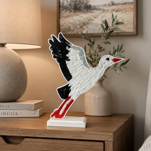 Könnte beinhalten: Eine Mosaikskulptur eines fliegenden Storchs aus weißen, schwarzen und roten Glasfliesen. Der Vogel ist mit ausgebreiteten Flügeln, einem spitzen roten Schnabel und langen roten Beinen dargestellt und auf einem rechteckigen weißen Sockel montiert.