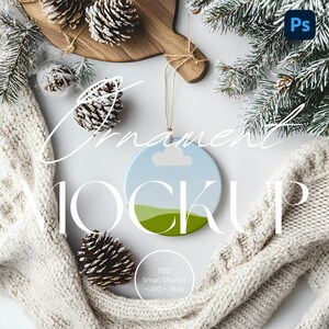 May include: A white round ornament with a blue sky and green hills design. The ornament is hanging from a string and is surrounded by pine branches, pinecones, and a white knitted scarf.