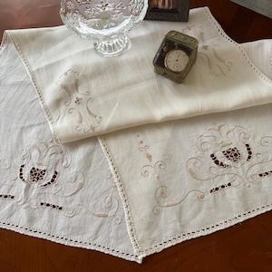 May include: An off-white rectangular table runner featuring intricate light brown embroidery with floral and scroll motifs, finished with a delicate crocheted lace border along the edges. The fabric has a smooth texture and a soft, neutral tone.