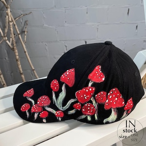 May include: Black baseball cap featuring a hand-painted design of red mushrooms with white spots. The mushrooms have green stems and leaves. The cap has a curved brim and a button on top. The text "IN STOCK" and "ONE SIZE" is printed on the side.