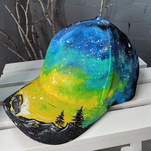 May include: A black baseball cap with a hand-painted design of a night sky with stars, a yellow and green aurora borealis, and a silhouette of a car driving through a mountain landscape with trees.