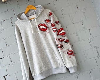 May include: A gray hoodie with a drawstring hood and a kangaroo pocket. The hoodie features a design of red lips on the sleeve and chest. The lips are painted with red lipstick and have a detailed, artistic style.