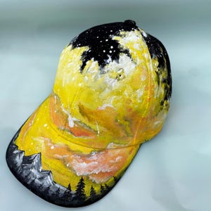 Puede incluir: Gorra de béisbol con un diseño de puesta de sol pintado. La gorra presenta una parte superior negra con motas blancas, que transita a un cielo amarillo y naranja con siluetas de montañas y árboles a lo largo del borde. Tamaño estándar.