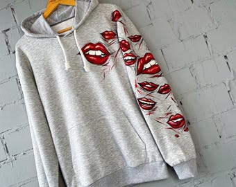 May include: A gray hoodie with a drawstring hood and a kangaroo pocket. The left sleeve and front of the hoodie feature a design of red lips. The lips are in various sizes and expressions, with red accents.