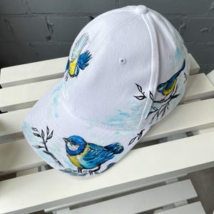 Könnte beinhalten: Eine weiße Baseballkappe mit handgemalten Illustrationen von blau-gelben Vögeln, die auf dünnen schwarzen Zweigen sitzen. Das Design umfasst zarte blaue Himmelsakzente und detaillierte Darstellungen der Vögel in verschiedenen Posen auf der Kappe.