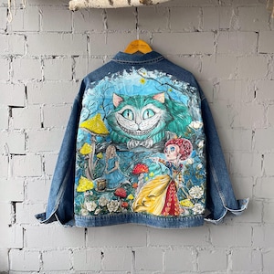 May include: A blue denim jacket featuring a hand-painted scene on the back. The artwork depicts a large green grinning cat, a small figure in a blue dress, and a figure in a yellow and red dress standing among yellow mushrooms and white flowers.
