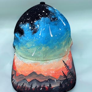 Könnte beinhalten: Baseballkappe mit handgemaltem Landschaftsdesign. Die Kappe zeigt einen Nachthimmel mit Sternen und Sternschnuppen über einer Bergkette und einem Wald. Die Farben sind Schwarz, Blau, Orange und Weiß.