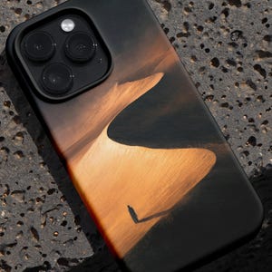 May include: Black smartphone case featuring a desert landscape design with a person silhouette. The case has a glossy finish and a raised camera module. The design showcases warm tones of orange and brown, with a dark background.