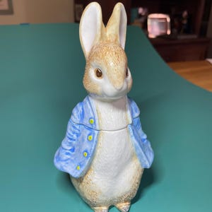 Puede incluir: Una figura de cerámica de un conejo de pie, con una textura de pelaje marrón claro y vistiendo una chaqueta azul brillante con botones amarillos. El conejo tiene orejas largas, ojos oscuros y pecho blanco, diseñado como un recipiente de dos piezas.