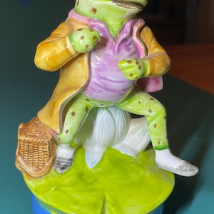 May include: A ceramic frog figurine wearing a yellow jacket and purple shirt, sitting on a white flower atop a green lily pad base. The frog has brown spots, orange eyes, and is positioned next to a small brown woven basket accessory.