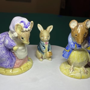 May include: Three ceramic animal figurines standing on yellow bases. On the left, a mouse in a purple dress and bonnet. In the center, a small rabbit in a blue jacket. On the right, a mouse in a blue coat and yellow vest.
