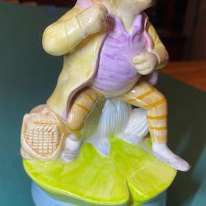 May include: A ceramic figurine of a frog wearing a yellow coat, a purple vest, and striped trousers. The frog sits on a green lily pad base with a small woven basket at its side. The figure features a glossy finish and detailed painted clothing.